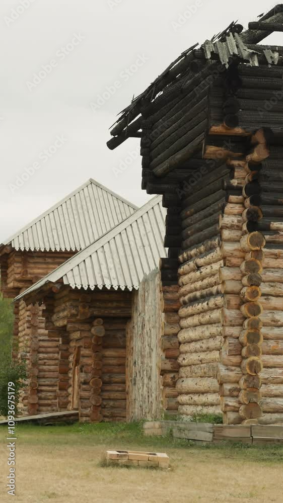 Old log house with burnt by fire roof on cloudy summer day. Medieval settlement with wooden buildings near birch grove. Ancient architectural objects Old log house with burnt by fire roof on cloudy summer day. Medieval settlement with wooden buildings near birch grove. Ancient architectural objects