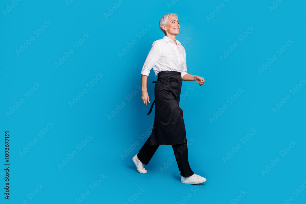 Full length photo of lovely pensioner lady walking serve clients coffee shop dressed waitress uniform isolated on blue color background Full length photo of lovely pensioner lady walking serve clients coffee shop dressed waitress uniform isolated on blue color background