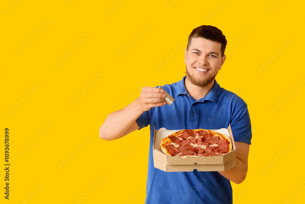 Young man holding cardboard box with tasty pepperoni pizza and salt on yellow background Young man holding cardboard box with tasty pepperoni pizza and salt on yellow background