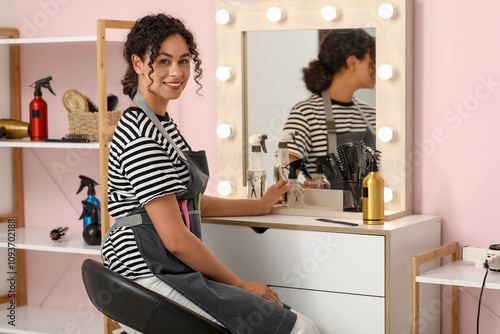 Beautiful young happy African-American hairdresser with professional tools sitting in beauty salon