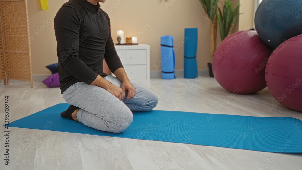 Naklejka premium Young hispanic man kneeling on a yoga mat in a gym surrounded by exercise equipment including fitness balls and dumbbells creating a serene workout atmosphere indoors