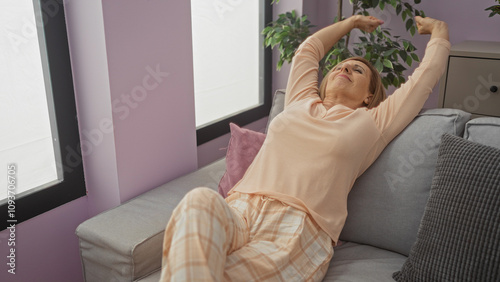 A mature caucasian woman relaxes in her home wearing pajamas, stretching comfortably on a sofa in a cozy living room filled with soft light and peaceful decor. photo