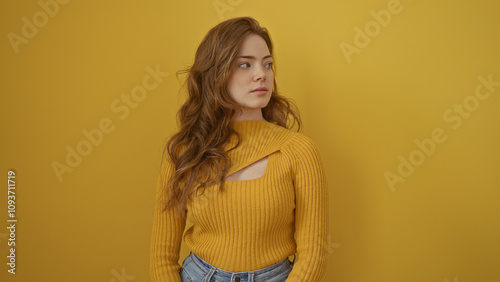 Attractive young woman with long blonde hair standing against an isolated yellow background wearing a fashionable mustard sweater.