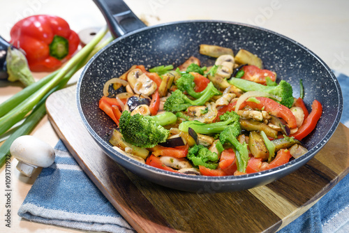 Colorful mixed vegetables in a frying pan such as broccoli, bell pepper, mushrooms, eggplant and onions on a wooden kitchen board, healthy vegetarian meal