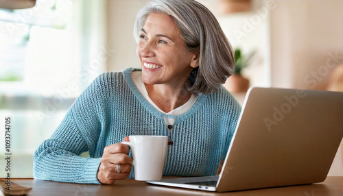 Happy mature woman sitting at home office table drinking coffee looking at laptop. Smiling relaxed middle aged older lady hybrid working on computer watching elearning webinar in modern house