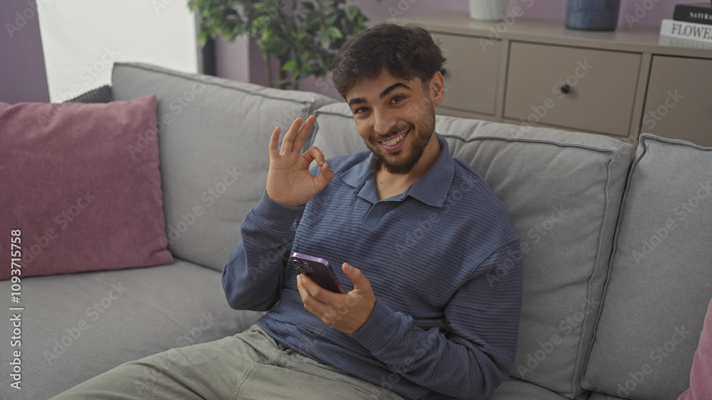 A cheerful man with a beard makes the okay gesture while holding a smartphone, sitting on a sofa in a cozy living room A cheerful man with a beard makes the okay gesture while holding a smartphone, sitting on a sofa in a cozy living room