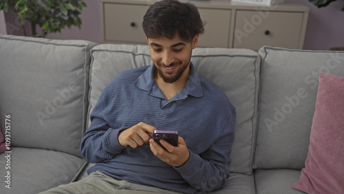 Handsome young man with beard using smartphone on grey couch indoors