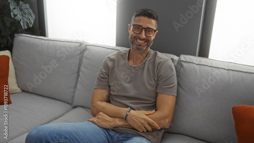 Handsome hispanic man smiles confidently while sitting indoors in a cozy home environment, capturing a sense of comfort and style in a modern living room setting.