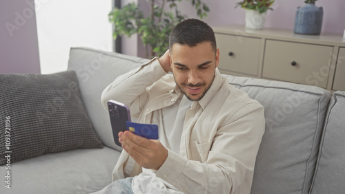 Handsome hispanic man in casual attire using smartphone on couch in modern apartment living room