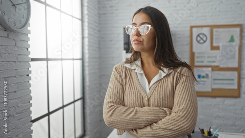 Fotografie Woman stands confidently in an office with arms crossed, wearing glasses, brunette hair, looking thoughtful, with a clock on the wall and sunlight streaming through windows