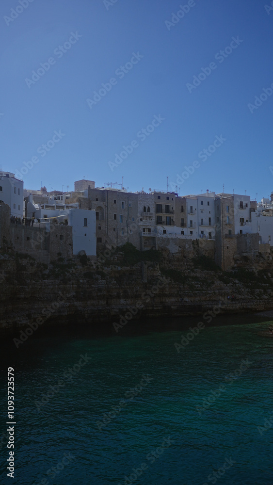 Naklejka premium Scenic view of the coastal town of polignano a mare in puglia, italy, showcasing its charming cliffside houses against a clear blue sky above the adriatic sea.