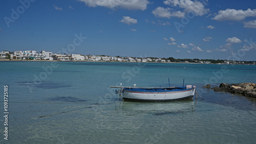 Colorful boat floating on turquoise water next to a rocky shore in porto cesareo, puglia, italy, with a backdrop of white buildings and a bright blue sky filled with fluffy clouds.