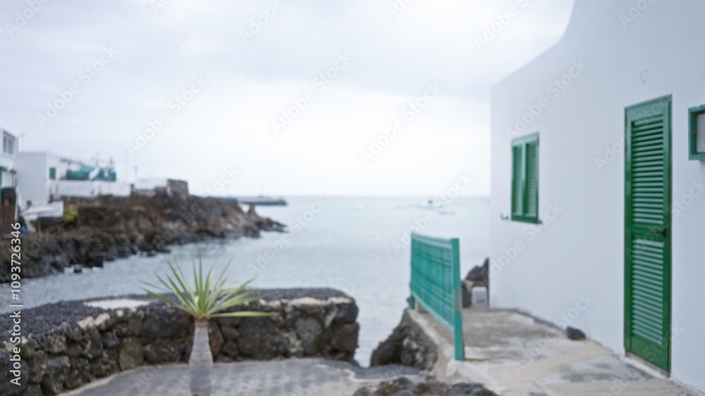 Fototapeta premium Blurred coastal scene in lanzarote with white buildings, green shutters, and rocky shoreline along the sea under a cloudy sky