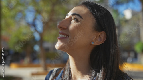 Obraz na plátně A smiling young hispanic woman enjoying the sun in a city park, with her denim jacket and earrings reflecting a casual urban vibe
