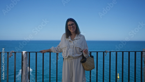 A young hispanic woman smiles beautifully while standing by a seaside railing in polignano a mare, puglia, italy, holding a straw bag against the stunning blue mediterranean backdrop.