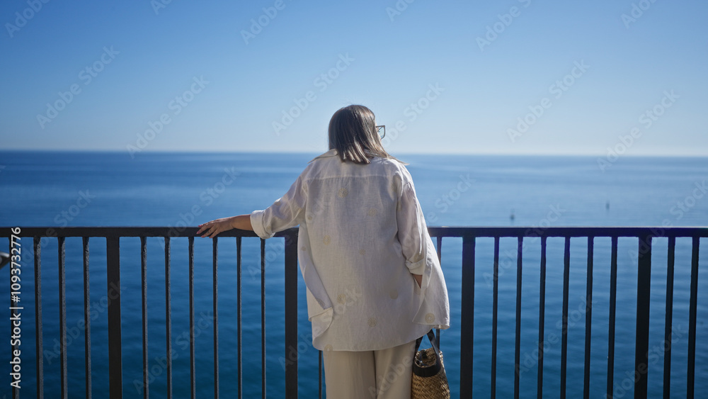 A young hispanic woman enjoys a serene moment at a viewpoint overlooking the ocean in polignano a mare, puglia, italy, with a backdrop of clear blue sky. A young hispanic woman enjoys a serene moment at a viewpoint overlooking the ocean in polignano a mare, puglia, italy, with a backdrop of clear blue sky.