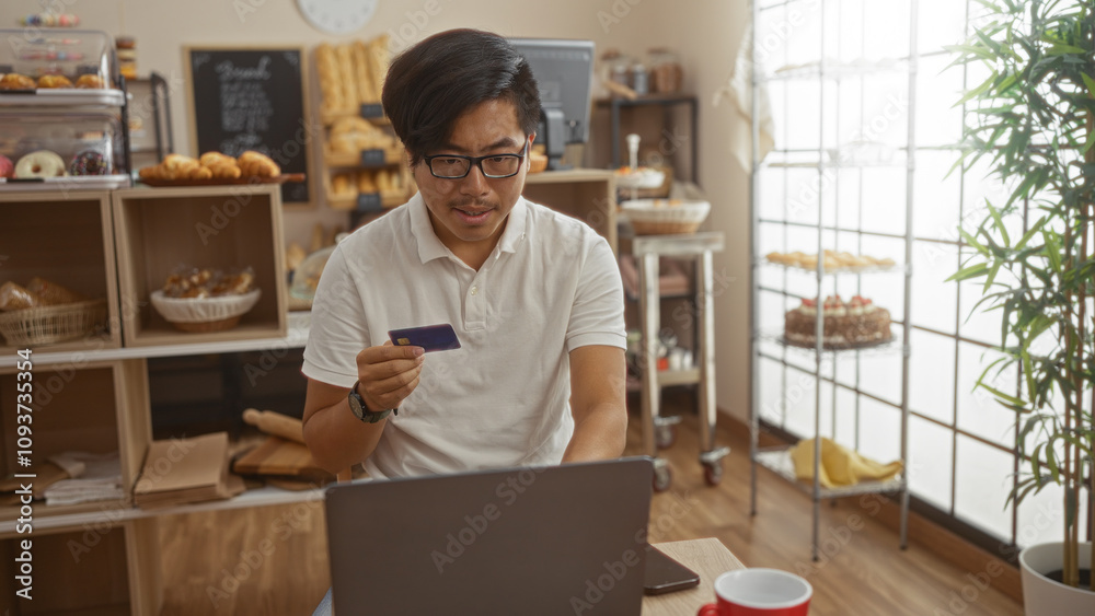 Young man in bakery using laptop and holding credit card with pastries on shelves in the background Young man in bakery using laptop and holding credit card with pastries on shelves in the background