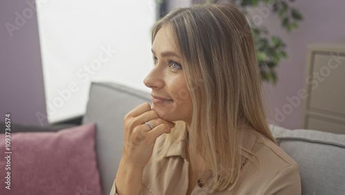 Beautiful blonde woman at home, sitting in a living room with a thoughtful expression, wearing casual attire and a ring, surrounded by soft pillows and cozy interior decor.