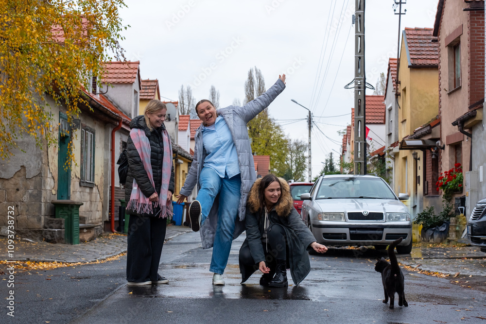 Obraz premium Three happy women enjoying moment with black cat on picturesque street lined with colorful houses and autumn trees. Concept of companionship, charm of small towns, and enjoying simple outdoor moments.