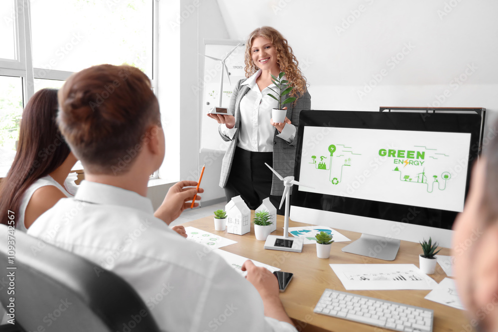 Obraz premium Young woman with wind turbine model giving presentation to her colleagues during business meeting in office. Green energy concept