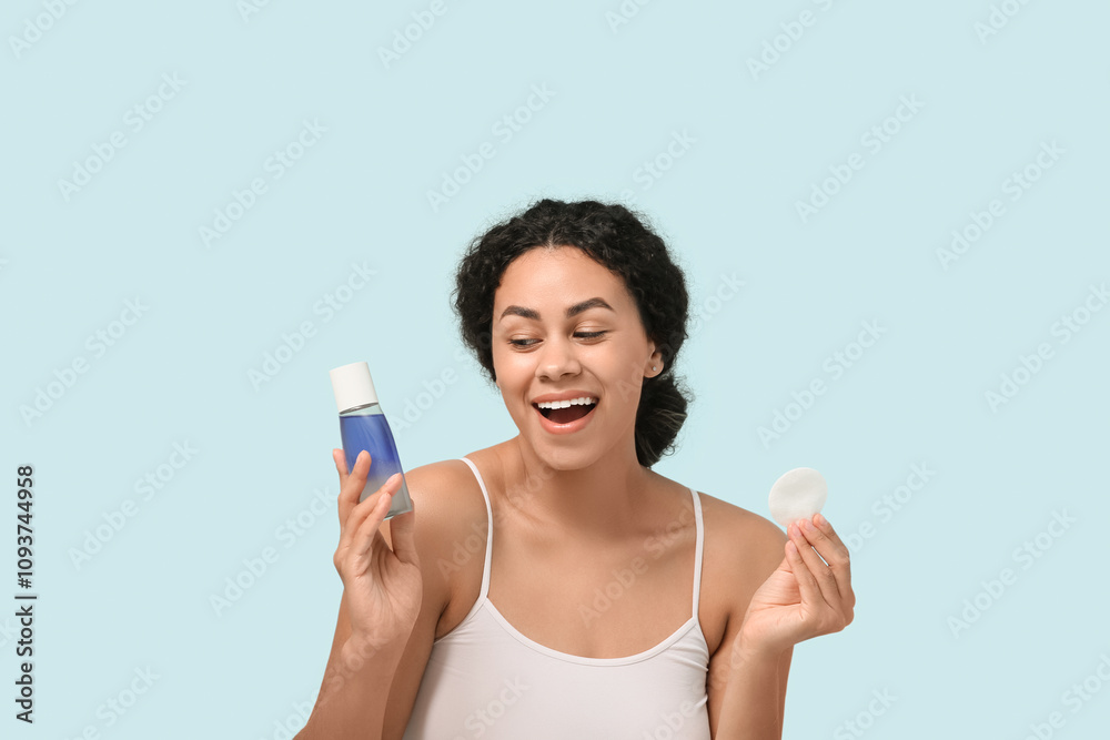 Happy young African-American woman with bottle of micellar water and cotton pad on blue background Happy young African-American woman with bottle of micellar water and cotton pad on blue background