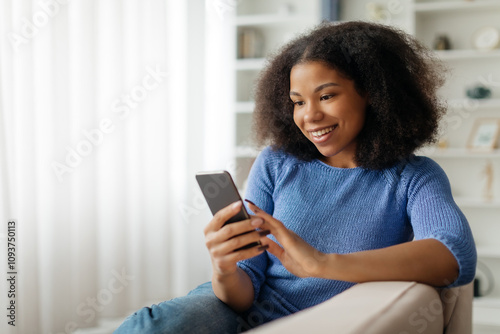 Portrait Of Happy Black Woman Using Mobile Phone At Home, Smiling Young African American Female Shopping Online On Smartphone Or Texting With Friends, Browsing New App While Relaxing On Couch. High