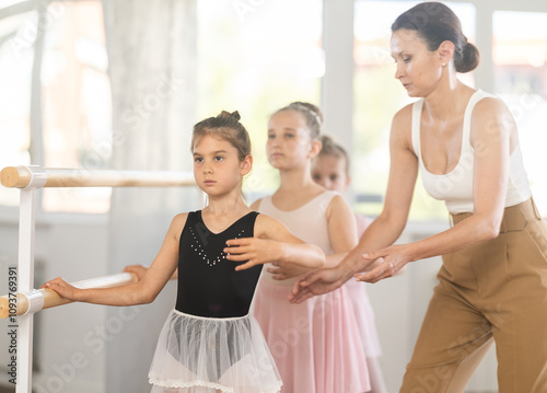 Adult woman teacher corrects group of girls dancers doing batman tandu movement at ballet barre photo