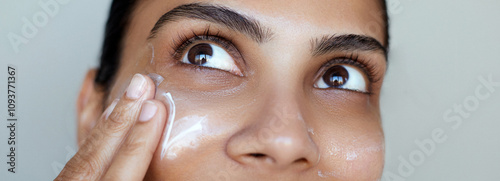 Beauty close-up south asian woman skincare portrait, one hand gently touching the face and applying a cream swatch or skin care cosmetic mask, facial hydration.