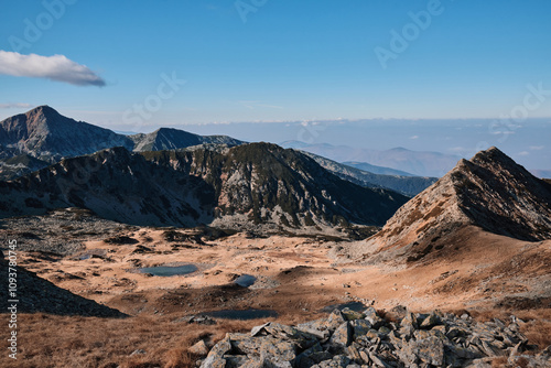 Stunning panoramic view of a mountain range under a clear blue sky. Rugged peaks, rocky terrain, and a tranquil lake create a breathtaking landscape, perfect for adventure and nature-themed projects.