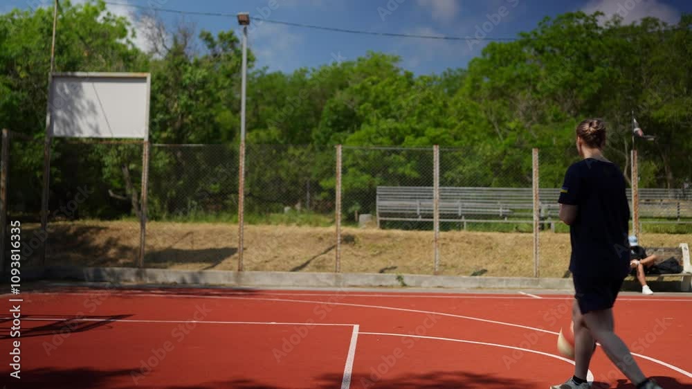 Side view of a happy blonde basketball player girl in a blue sports uniform warming up and hitting the ball off the floor during basketball training on a street sports ground in the summer Side view of a happy blonde basketball player girl in a blue sports uniform warming up and hitting the ball off the floor during basketball training on a street sports ground in the summer