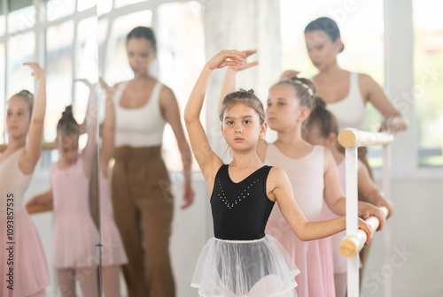 Adult woman teacher helping group of girls dancers practicing batman tandu movement at barre in studio photo