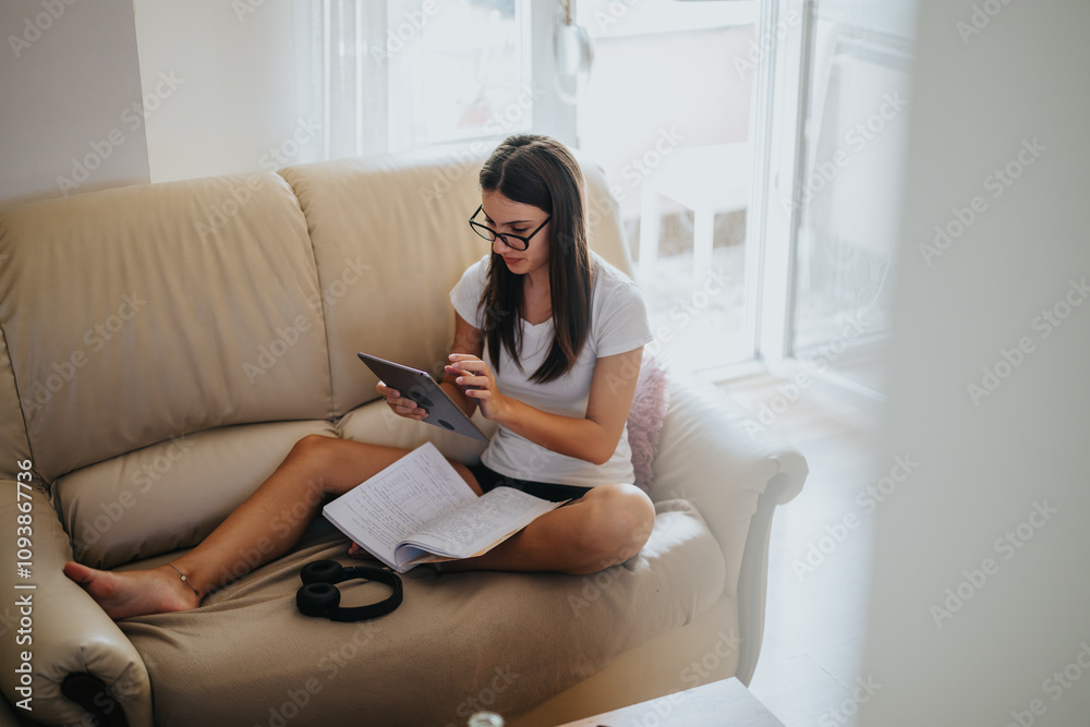 A young woman sits on a sofa, studying with a tablet and notebook. This cozy home setting reflects focus, relaxation, and modern remote education. A young woman sits on a sofa, studying with a tablet and notebook. This cozy home setting reflects focus, relaxation, and modern remote education.