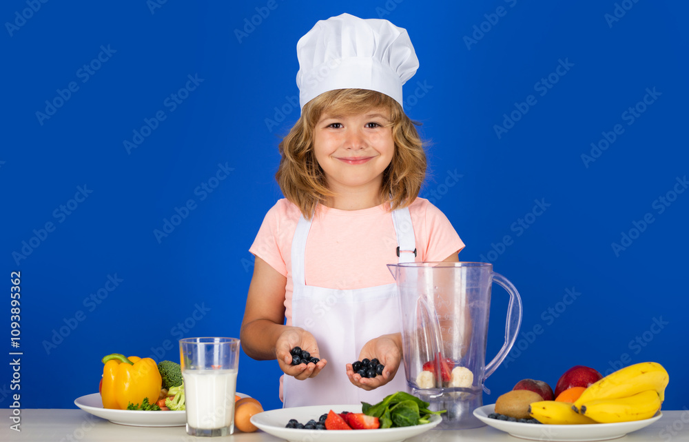 Child chef cook prepares food in isolated blue studio background. Kids cooking blueberries smoothie. Teen boy with apron and chef hat preparing a healthy vegetables meal in the kitchen. Child chef cook prepares food in isolated blue studio background. Kids cooking blueberries smoothie. Teen boy with apron and chef hat preparing a healthy vegetables meal in the kitchen.