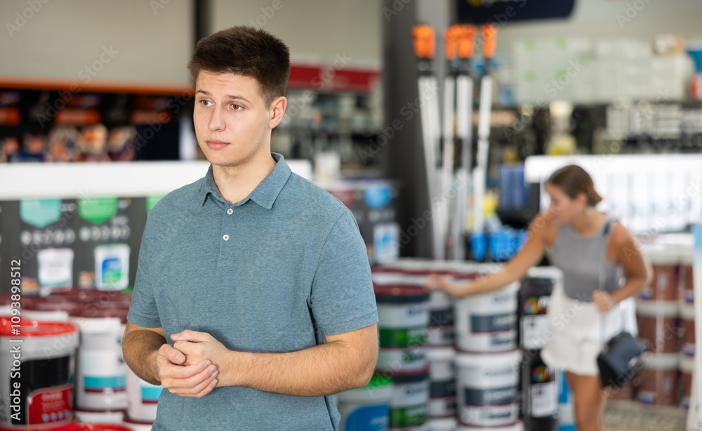 Young man in trading hall of store of construction goods paints and varnishes stand and remembers goods necessary for purchase Young man in trading hall of store of construction goods paints and varnishes stand and remembers goods necessary for purchase