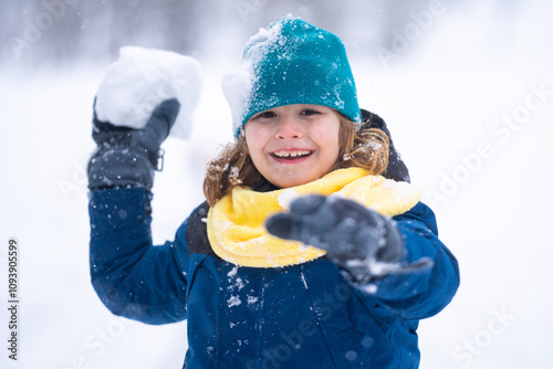 Happy Child playing with snow during snowfall. Snowball fight Child in winter clothes throw snowball. Winter children play with snow. Kid play with snowball. Winter kids face. Boy play snowball. photo