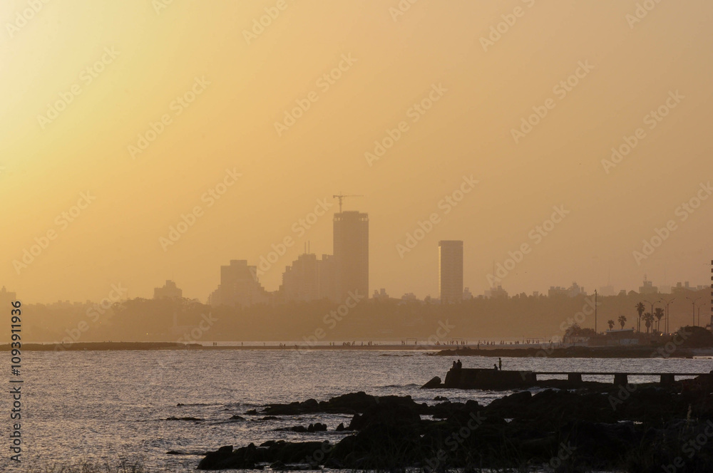 Sunset skyline silhouette over calm sea and rocky shore, tranquil urban scene Sunset skyline silhouette over calm sea and rocky shore, tranquil urban scene