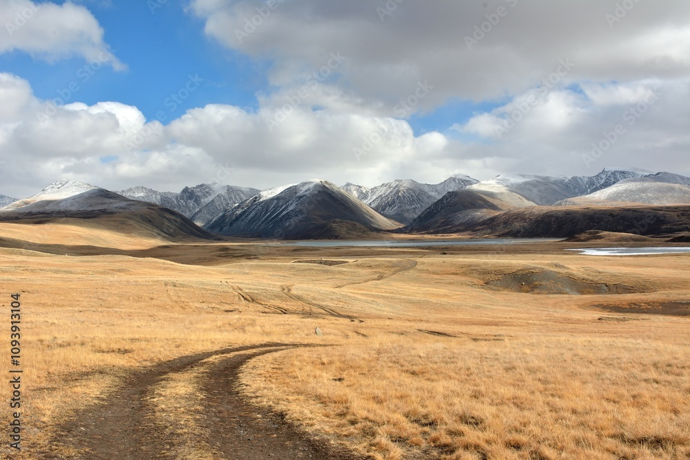 The field road, turning to the right, descends from a high hill into a picturesque valley against the backdrop of snow-capped mountain peaks. The field road, turning to the right, descends from a high hill into a picturesque valley against the backdrop of snow-capped mountain peaks.