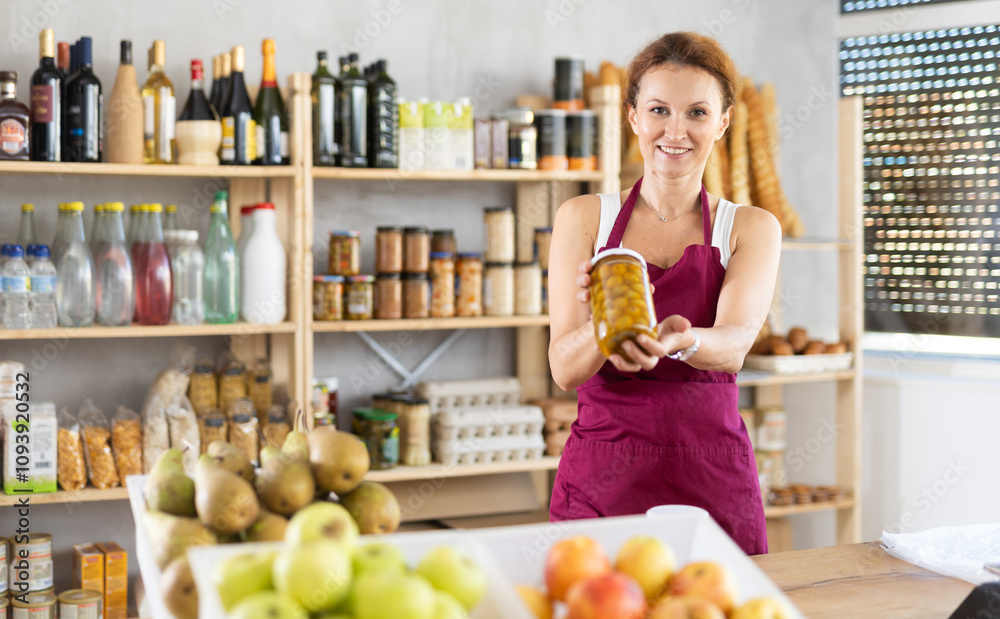 Adult female seller at counter offering olives in jar in grocery store Adult female seller at counter offering olives in jar in grocery store