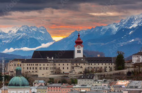 A striking sunset view of Nonnberg Abbey in Salzburg, Austria, with its iconic red clock tower set against the backdrop of snow-capped mountains and a dramatic cloudy sky photo