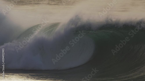 Large wave barrels forward as mist trails off the crest, surfers visible in lineup photo