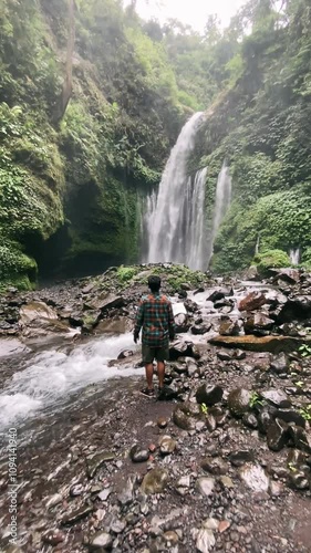 Man walks towards a beautiful waterfall on Mount Rinjani with a forest of tropical green trees at Tiu Kelep Waterfall, Lombok, Indonesia. 