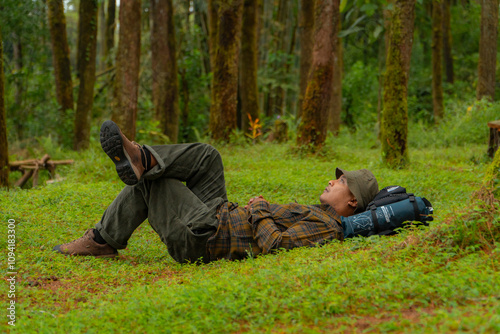 An adventurer was lying on the green grass in a beautiful and quiet pine forest area. Male travelers enjoy the serenity of the wilderness by lying down on the green grass in the pine forest