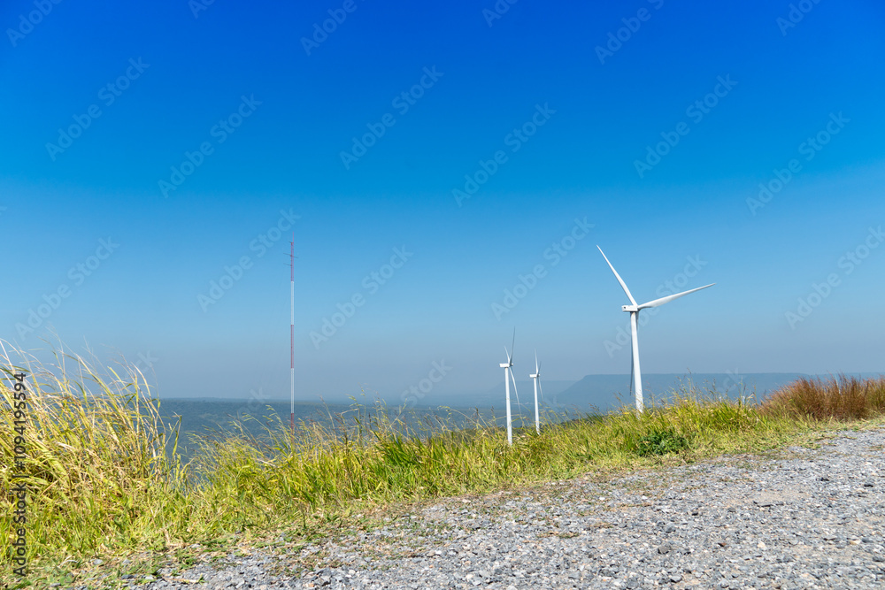 Viewpoint with gravel and grass growing on the green edge. Wind turbines tower over a lush forest. Background of mountain far away under blue sky. Landmark of Khao Yai Thieng. Viewpoint with gravel and grass growing on the green edge. Wind turbines tower over a lush forest. Background of mountain far away under blue sky. Landmark of Khao Yai Thieng.