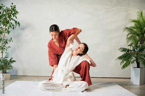 Masseuse making thai yoga massage. Woman therapist doing traditional massage treatment, stretching female patient's body. Client sitting on mat, enjoying hand massage in calm atmosphere. photo