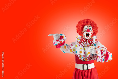 Surprised clown pointing at blank template copy spase for advertising, posters, greetings, invitations. Entertainer as Joker in a suit and wig, with clown whiteface makeup photo