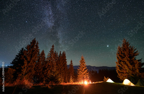 Panoramic view of mountain hill with illuminated camp tents, coniferous trees and hikers under starry sky. Night camping in mountains under beautiful sky with stars. Concept of hiking and traveling. photo