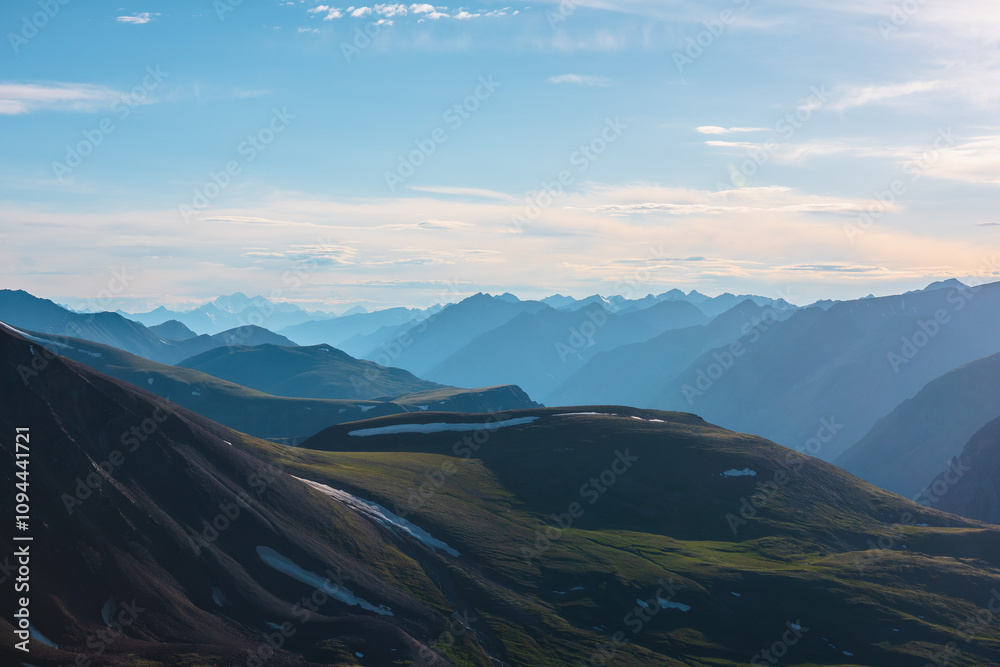Aerial top view to big green hills and large rocky mountains silhouettes in bright sun under clouds in blue sky. Sunlit high snowy mountain range far away. Scenic layered alpine landscape at sunset. Aerial top view to big green hills and large rocky mountains silhouettes in bright sun under clouds in blue sky. Sunlit high snowy mountain range far away. Scenic layered alpine landscape at sunset.