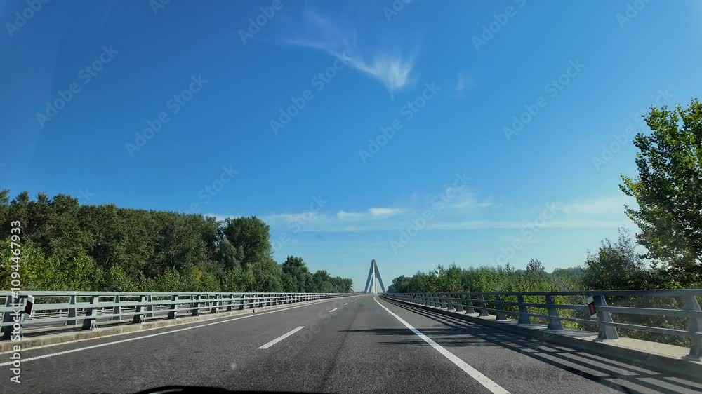 A car drives along a Croatian road under clear skies, approaching a bridge flanked by green trees. The scene captures the smooth drive across the modern bridge surrounded by scenic nature. A car drives along a Croatian road under clear skies, approaching a bridge flanked by green trees. The scene captures the smooth drive across the modern bridge surrounded by scenic nature.
