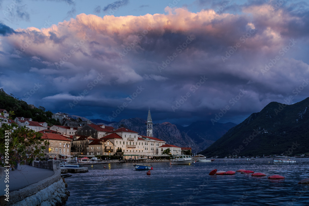 The view оf Perast, Montenegro, illuminated by the warm, golden light of the setting sun. The foreground is serene, with boats floating, while dark storm clouds loom dramatically on background The view оf Perast, Montenegro, illuminated by the warm, golden light of the setting sun. The foreground is serene, with boats floating, while dark storm clouds loom dramatically on background