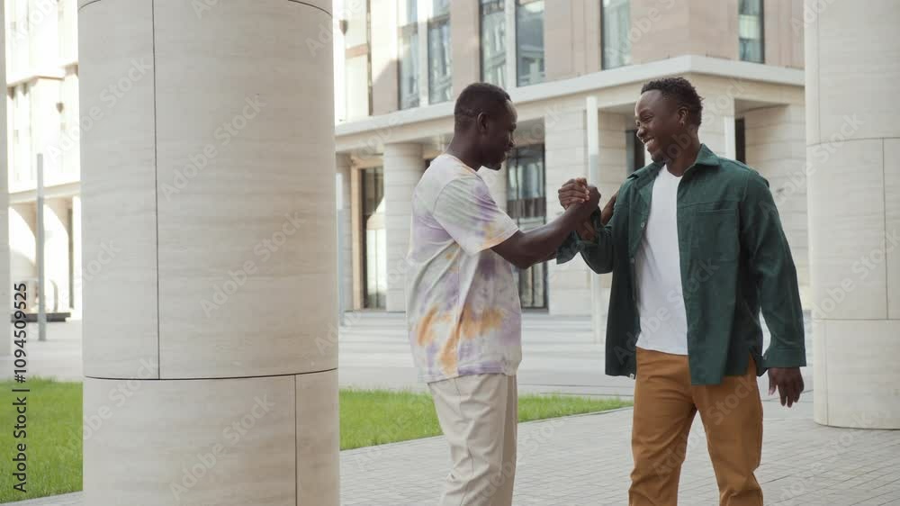 Two African American male students stand near buildings, talking. Joyful young man co workers on break chill outdoors chatting. Cheerful black skinned friends shake hands, walk away saying goodbye Two African American male students stand near buildings, talking. Joyful young man co workers on break chill outdoors chatting. Cheerful black skinned friends shake hands, walk away saying goodbye